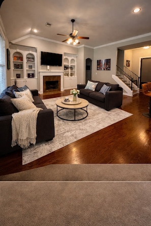 A living room with a fireplace, a ceiling fan, a staircase, and a TV mounted on the wall.