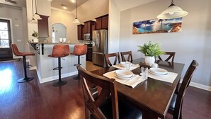 A dining area with a wooden table, four chairs, a chandelier, and a kitchen in the background.