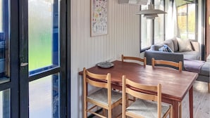 Un coin repas avec une table et des chaises en bois, une estampe botanique encadrée au mur et une fenêtre donnant sur un paysage verdoyant.