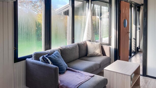 Un salon moderne avec un canapé d’angle gris, une petite table d’appoint en bois et de grandes fenêtres donnant sur l’extérieur.