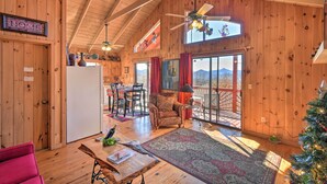 Una habitación amplia con paredes de madera, un ventilador de techo, un comedor con sillas y una mesa, un ventanal con cortinas y un árbol de Navidad.