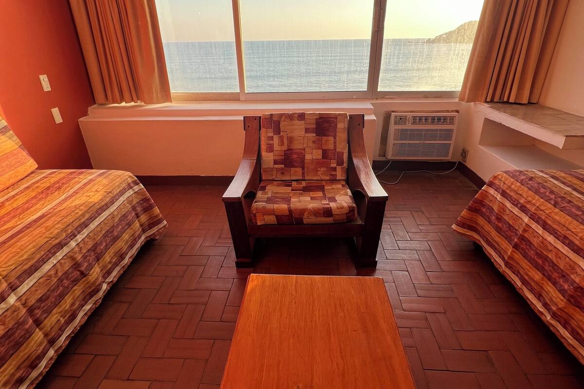 Une chambre avec deux lits, une table en bois, une chaise avec un dossier en bois et une grande fenêtre donnant sur la mer.