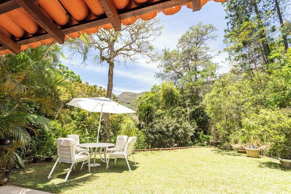An outdoor patio with a table and chairs under a pergola, surrounded by lush greenery and a mountain backdrop. - Casa com Piscina Churrasqueira Jogos e Lareira (Petrópolis)