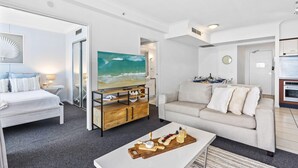 Une chambre d’hôtel moderne équipée d’un lit, d’un canapé, d’une table basse avec des collations, d’une télévision et d’un tableau représentant une scène de plage.