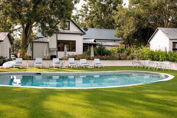 Une piscine avec des chaises longues, une maison équipée d’une antenne parabolique et un jardin avec différentes espèces de plantes.
