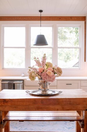 Une table en bois surmontée d’un vase de fleurs, dans une cuisine dotée de grandes fenêtres et d’un luminaire suspendu.
