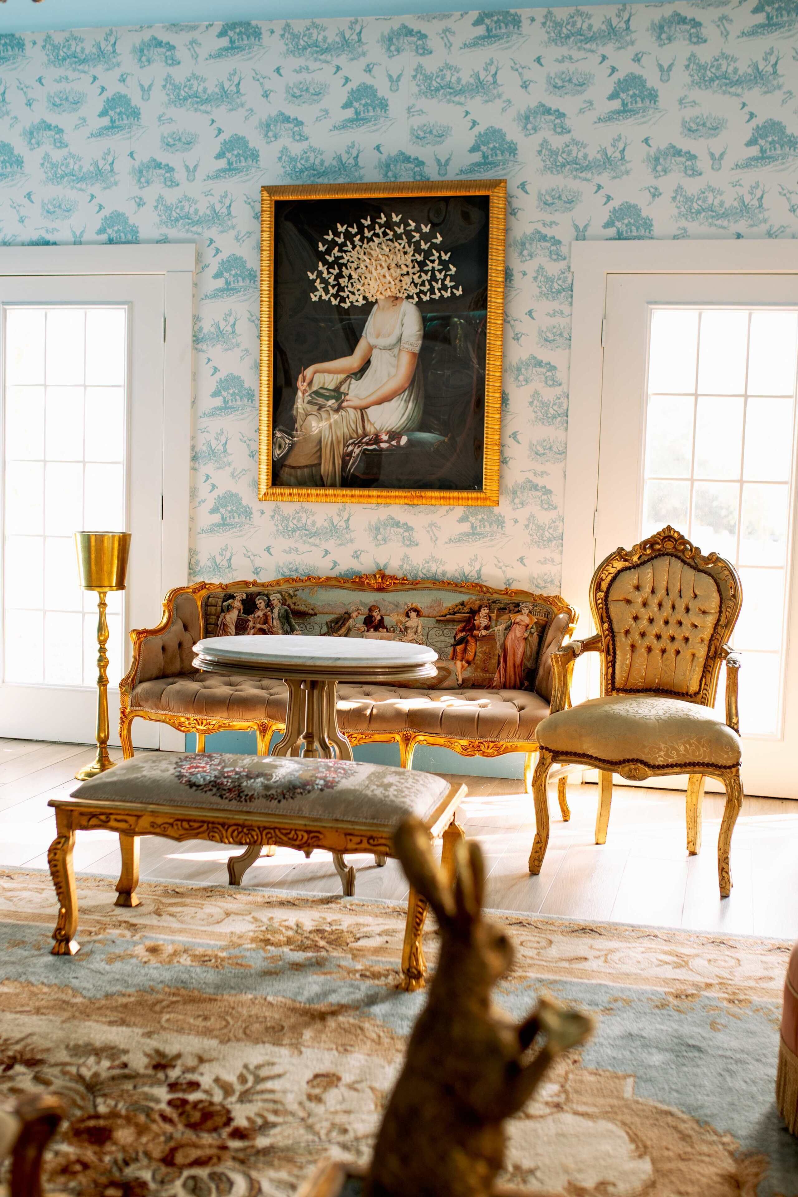 Une pièce comprenant un canapé, un fauteuil et un pouf, un tableau au mur et un papier peint à motifs.