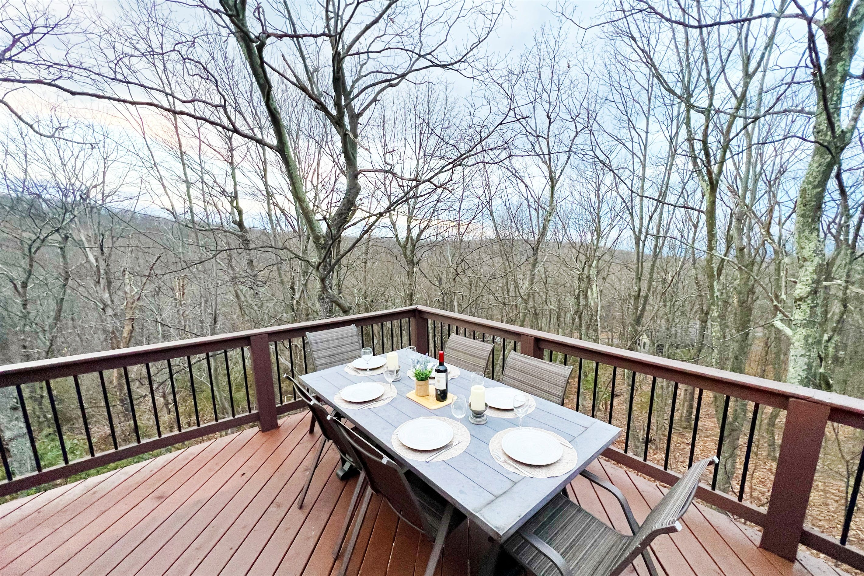 A wooden deck with a table set for four, surrounded by chairs, overlooking a forested area.