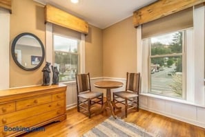 Quarto nos fundos com cômoda de madeira, mesa redonda com cadeiras, espelho e janelas amplas com vista para a rua.