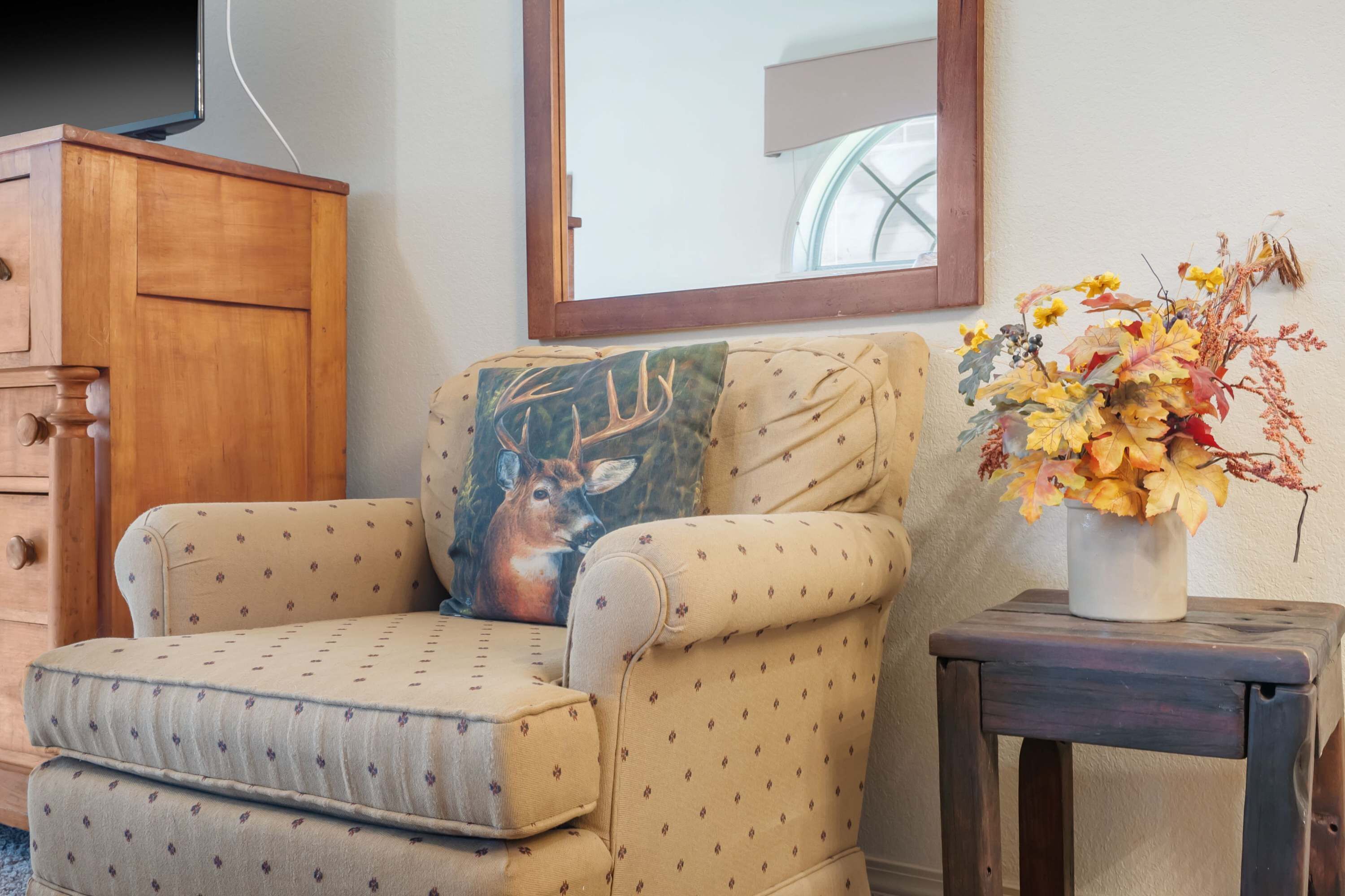 A beige armchair with a deer pillow, a small wooden table with a vase of autumn leaves, and a wooden cabinet.