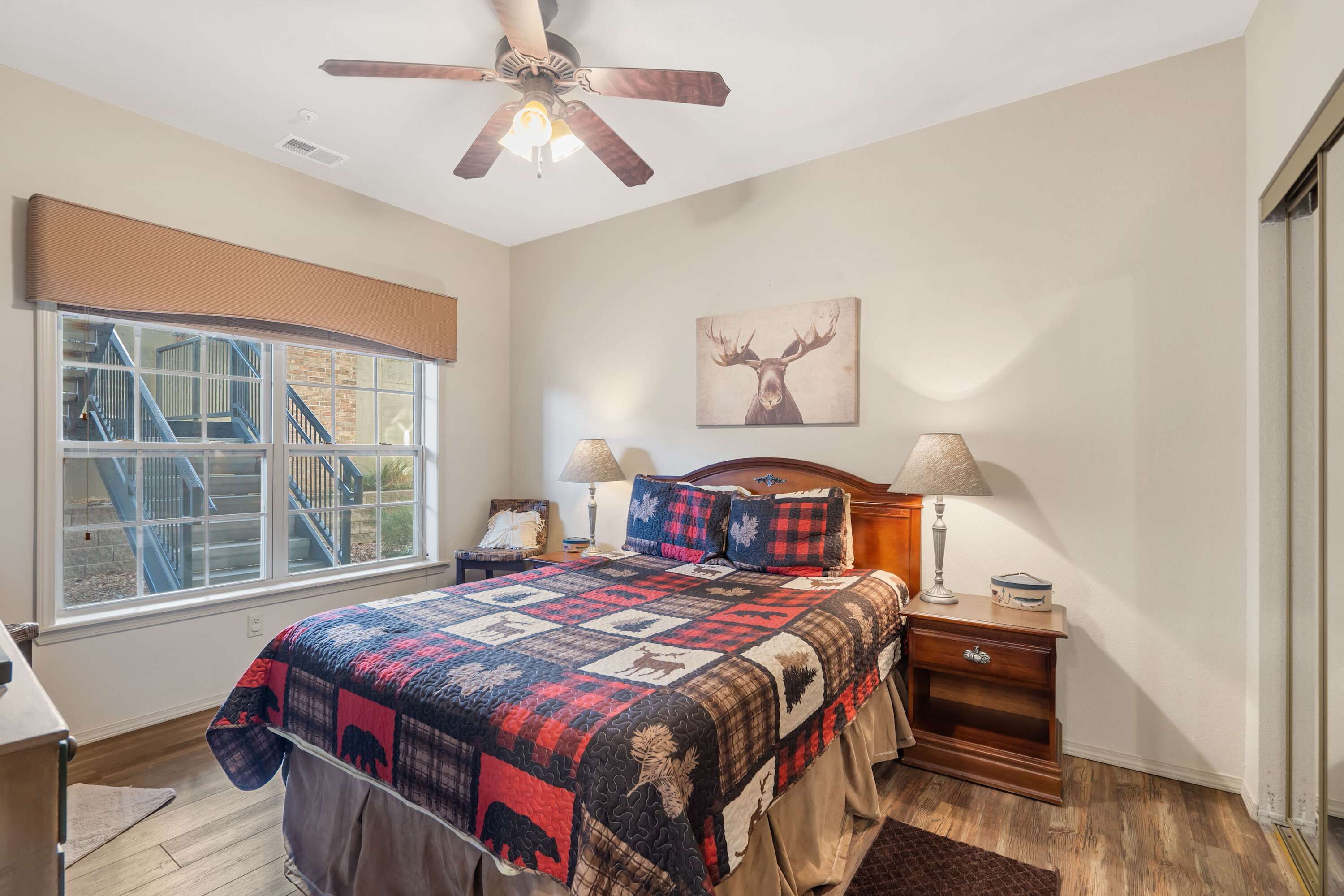 A bedroom with a large bed, a ceiling fan, a window with a view of a staircase, and a deer painting on the wall.