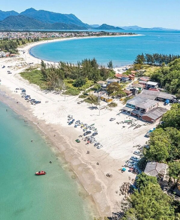 An aerial view of a coastal area with a beach, a small boat, and buildings. - Ampla Casa com Piscina Ponta do Papagaio (Palhoça)