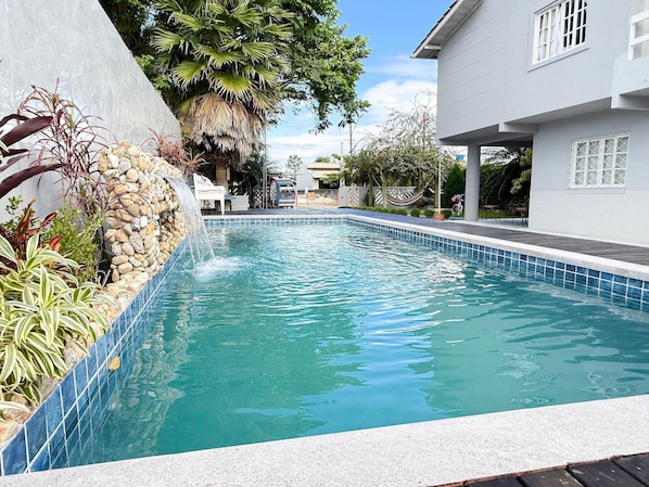 A swimming pool with a stone waterfall feature, surrounded by tropical plants and a modern house.