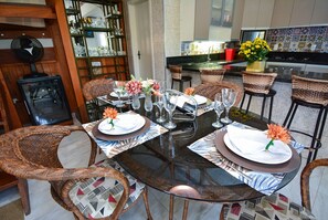 Un comedor con mesa de vidrio, sillas de mimbre y una zona de bar con estantes y un cubo rojo.