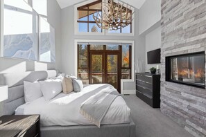 Ein modernes Schlafzimmer mit einem großen Bett, einem Kamin, einem Fernseher und Blick auf Herbstbäume durch die Fenster.