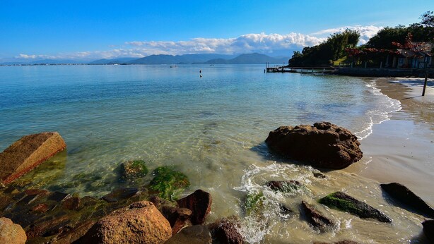 A beach with clear water, rocks, and a distant mountain range. - Casa à Beira Mar no Ribeirão -SDI012 (Florianópolis)