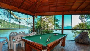 A pool table with a green felt surface, surrounded by white plastic chairs, in a room with large windows overlooking a body of water and greenery. - Casa à Beira Mar no Ribeirão -SDI012 (Florianópolis)