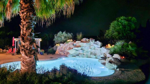 Un espace piscine aménagé pour la nuit, avec une cascade en pierre, entouré d’une végétation tropicale et d’un palmier.
