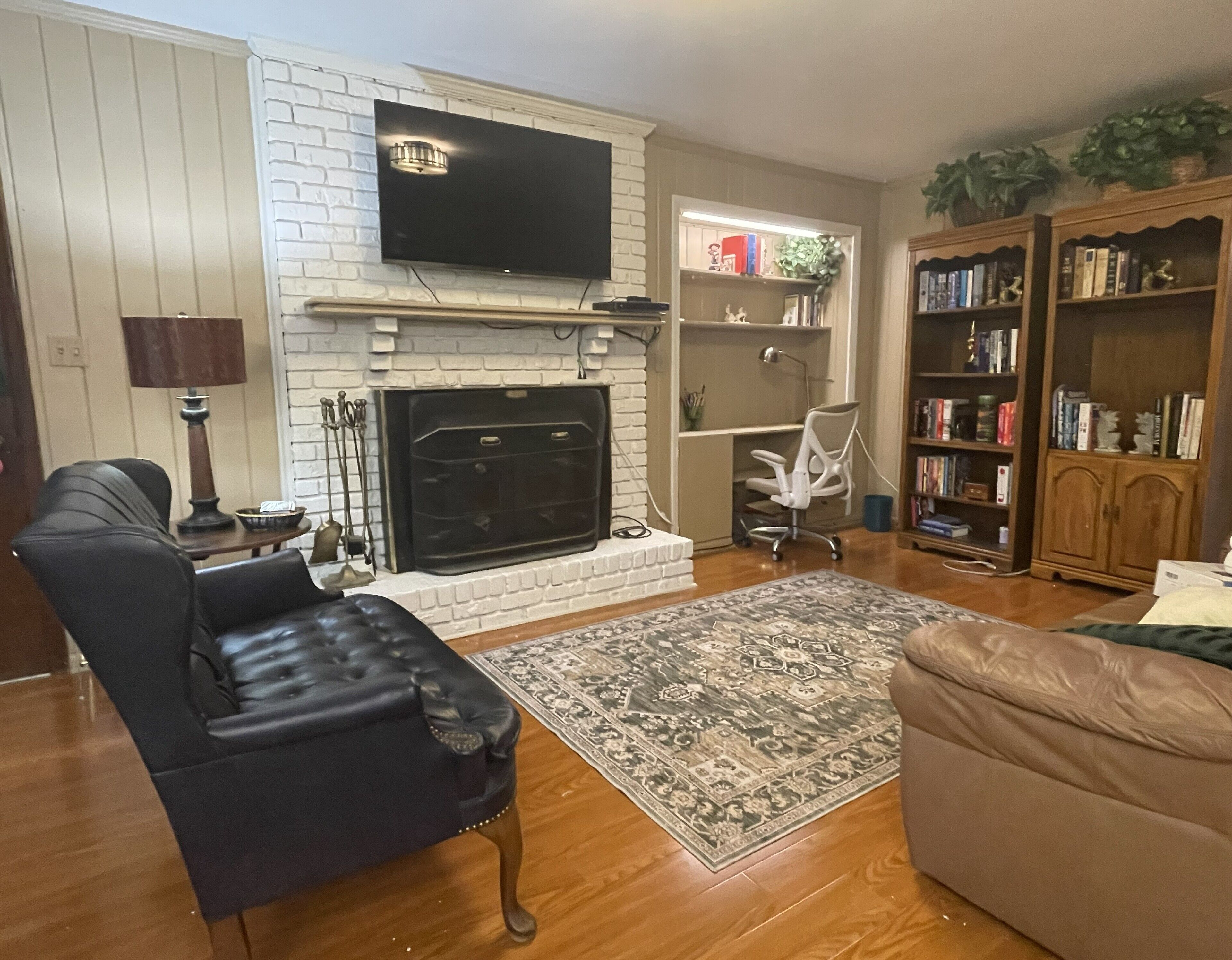 A living room with a fireplace, a television mounted above it, a leather armchair, a beige sofa, a patterned rug, and a bookshelf filled with books.