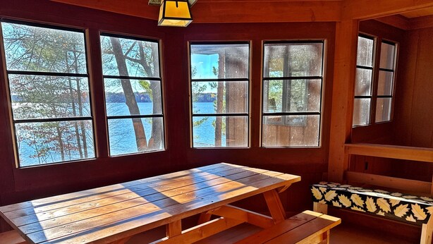 A wooden table and benches in a room with large windows overlooking a body of water and trees.