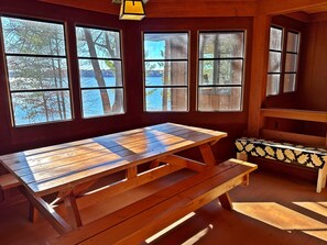 Huoneessa on puinen pöytä ja penkit, ja suurista ikkunoista avautuu näkymä vesistöön ja puihin.