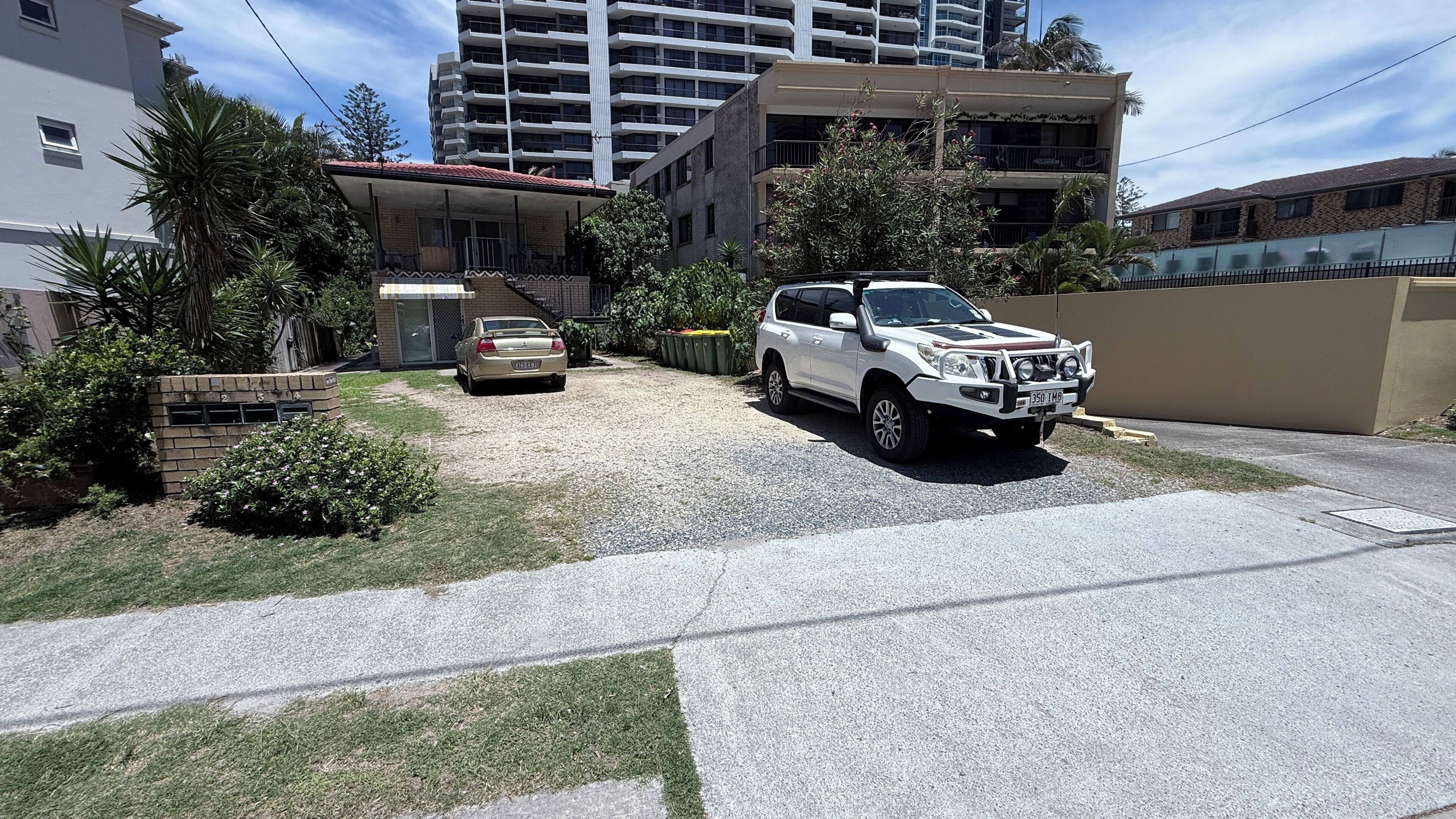 Un quartier résidentiel avec un VUS blanc garé devant une maison, une voiture beige sur l’allée, et un grand immeuble en arrière-plan.