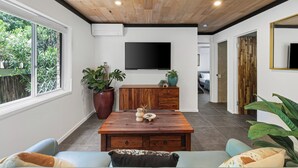 Un salon moderne avec un plafond en bois, un grand téléviseur fixé au mur, un meuble en bois et une table à manger agrémentée de quelques objets décoratifs.