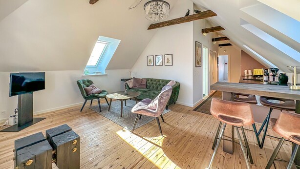 Et loft-agtigt opholdsrum med højt til loftet, trægulv, en grøn veloursofa, et sofabord og et barområde med barstole.