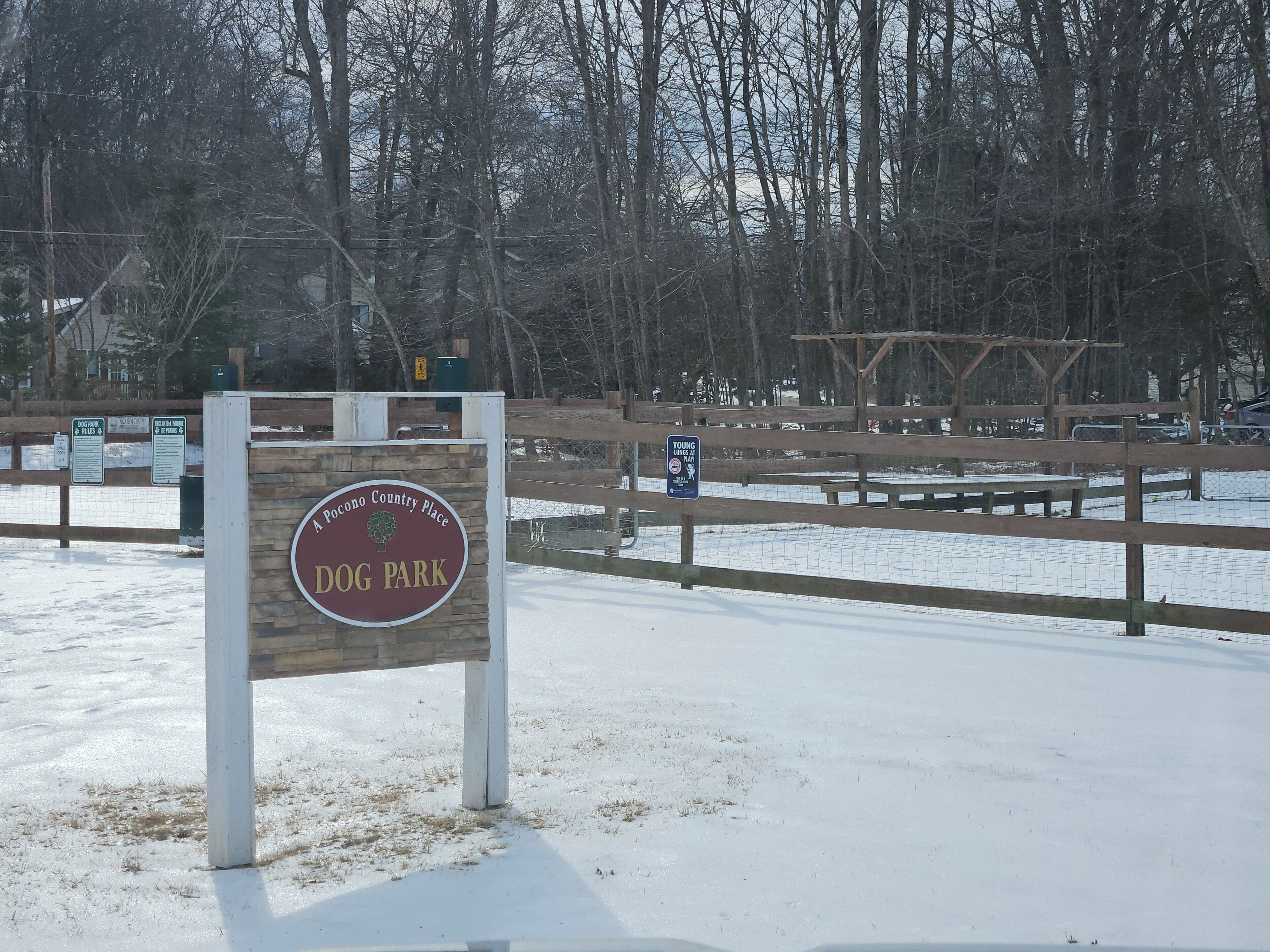 Schild für den Hundepark mit Holzzaun und Bäumen im Hintergrund.