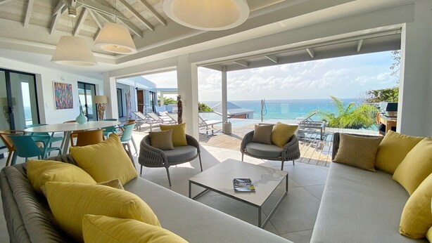 Ein modernes Wohnzimmer mit einer großen Couch, Sesseln und einem Couchtisch. Blick auf den Ozean und ein Pool sind von außen zu sehen.