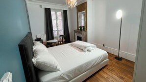A bedroom with a bed, a bedside table, a chair, a small table, a mirror, a fireplace, and a floor lamp. - Bright & Cozy Paris Stay in Montparnasse (Paris)