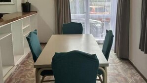 Quarto com uma mesa branca e quatro cadeiras, uma janela com cortinas, um radiador e um pequeno armário com um rádio.
