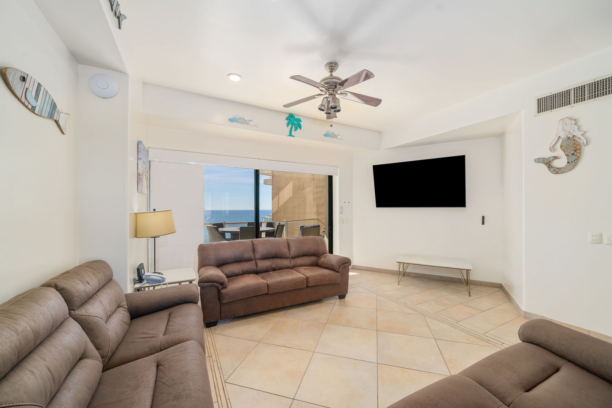 A spacious living area with a brown sofa, a ceiling fan, a flat-screen TV, and decorative wall art.
