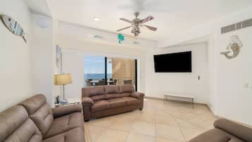 A spacious living area with a brown sofa, a ceiling fan, a flat-screen TV, and decorative wall art.