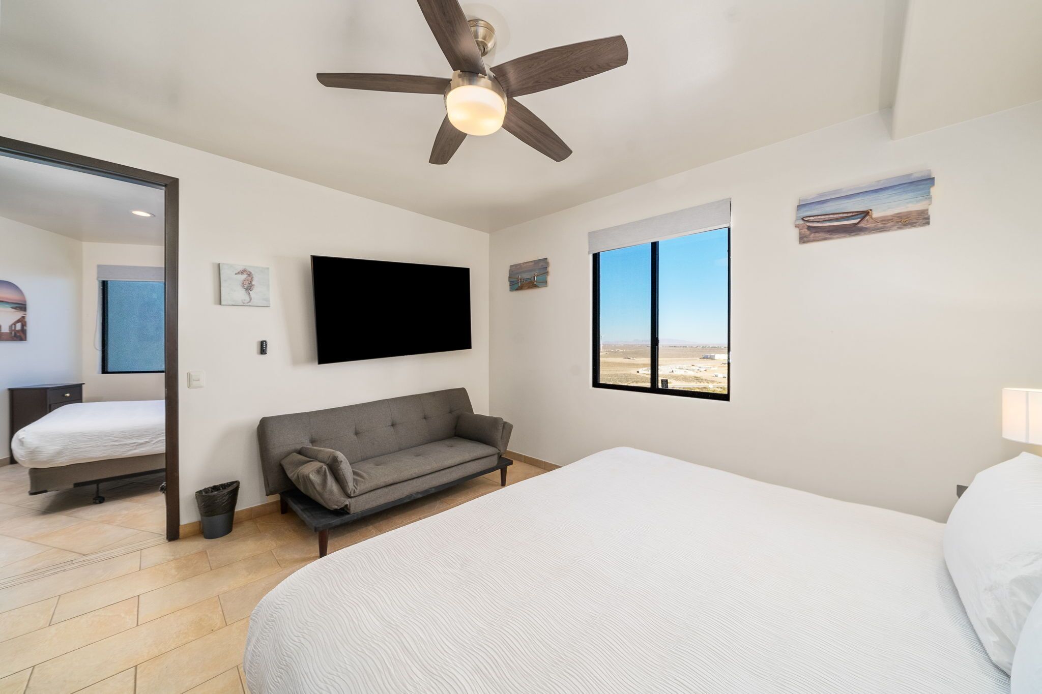 A bedroom with a bed, a gray sofa, a ceiling fan, a TV, and a window with a desert view.