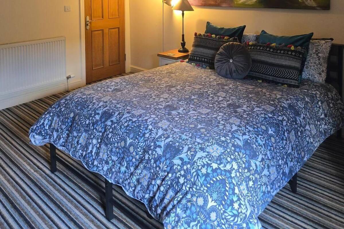 A bedroom with a large bed, a nightstand, a lamp, a door, and a colorful abstract painting on the wall.