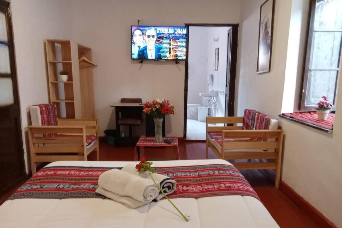 Quarto de hotel com cama, duas cadeiras de madeira, uma mesinha com um vaso de flores, televisão na parede e janela com vista.