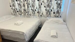 Two single beds with white linens and rolled towels, a patterned curtain, and a carpeted floor. - 4 Beds Including 1 Sofa Bed in 1 Bedroom+1 Lounge (Auckland)