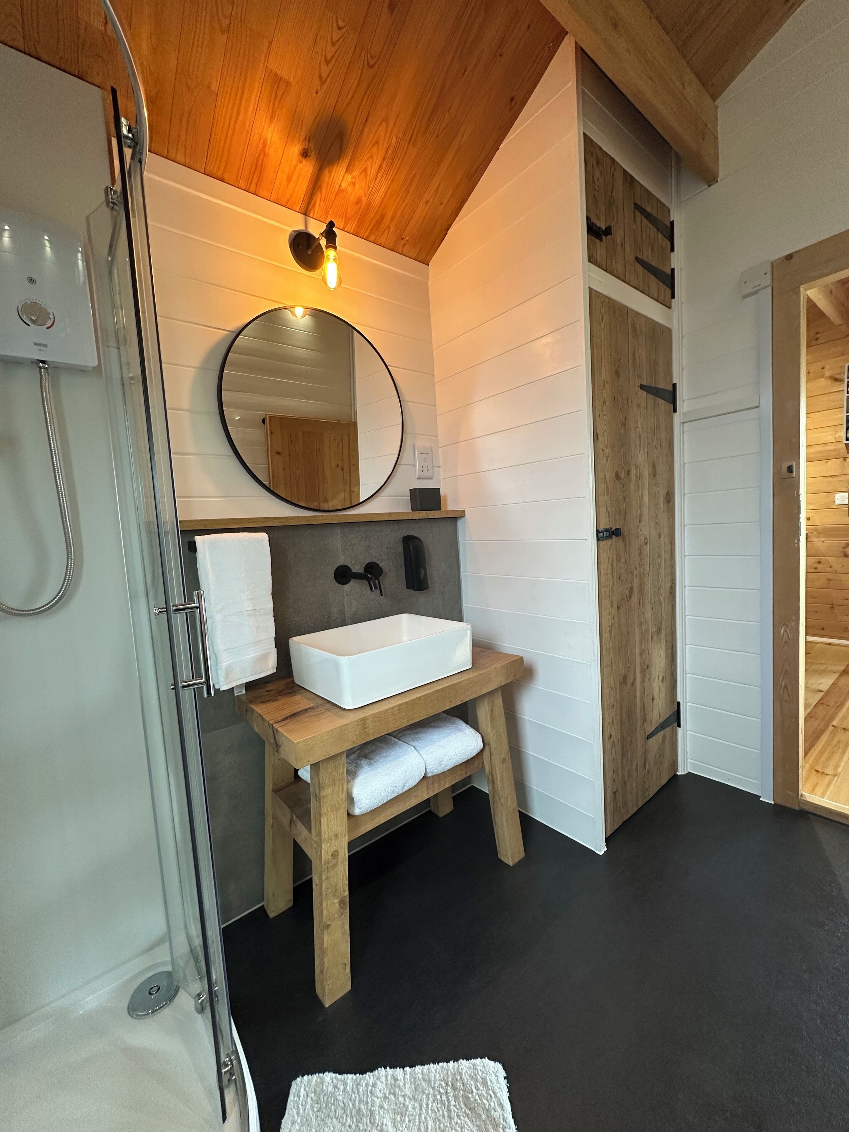 Un baño con tocador de madera, un lavamanos blanco, un espejo redondo y una ducha.