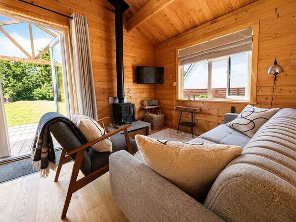 Et hyggeligt sommerhusinteriør med sofa, fjernsyn, pejs og et vindue med udsigt til en have.
