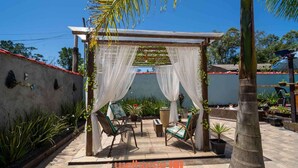 A covered outdoor seating area with chairs and a table, surrounded by potted plants and a palm tree. - Sítio em Mongaguá com Piscina Grande e Lazer (Mongaguá)