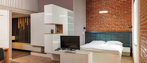 A modern bedroom with a brick wall, a bed with white bedding, a TV on a white cabinet, and a wooden floor.