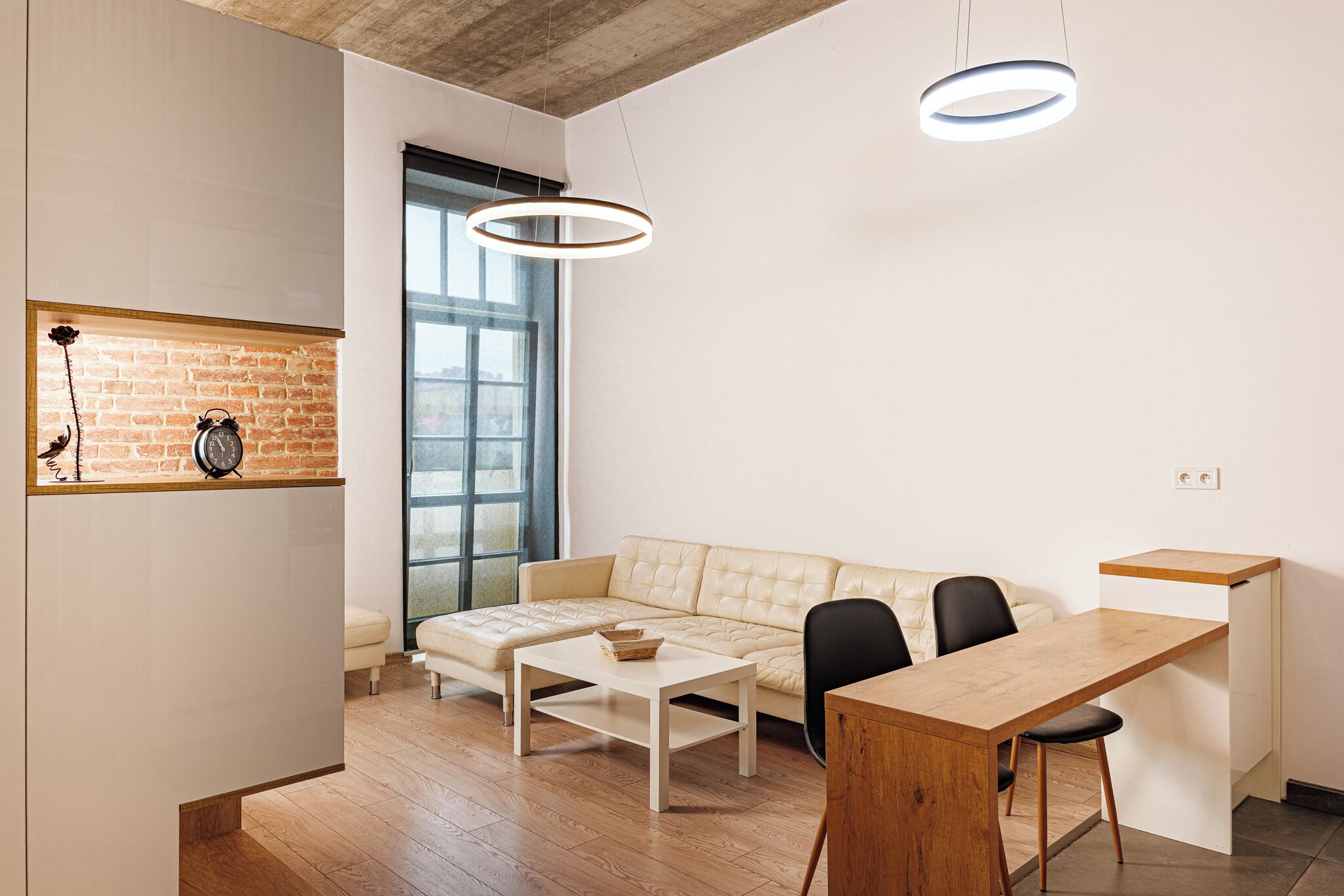 Un espacio habitable moderno con un sofá, una mesa de centro, una mesa de comedor y dos sillas. Se aprecia una pared de ladrillo y un ventanal grande que proporciona luz natural.