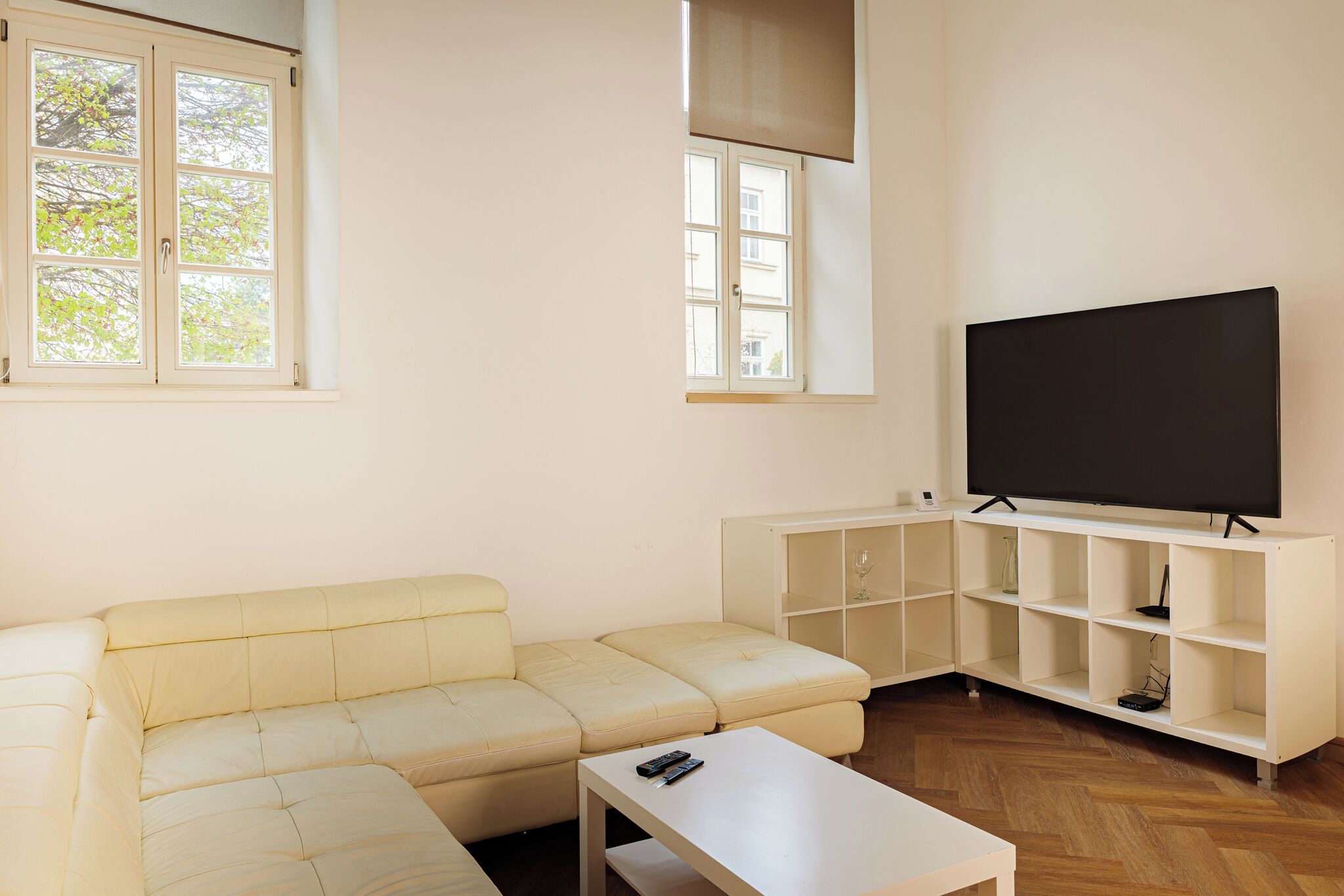 Una sala de estar con un sofá color beige, una mesa de centro blanca, un televisor sobre un mueble blanco y piso de madera.