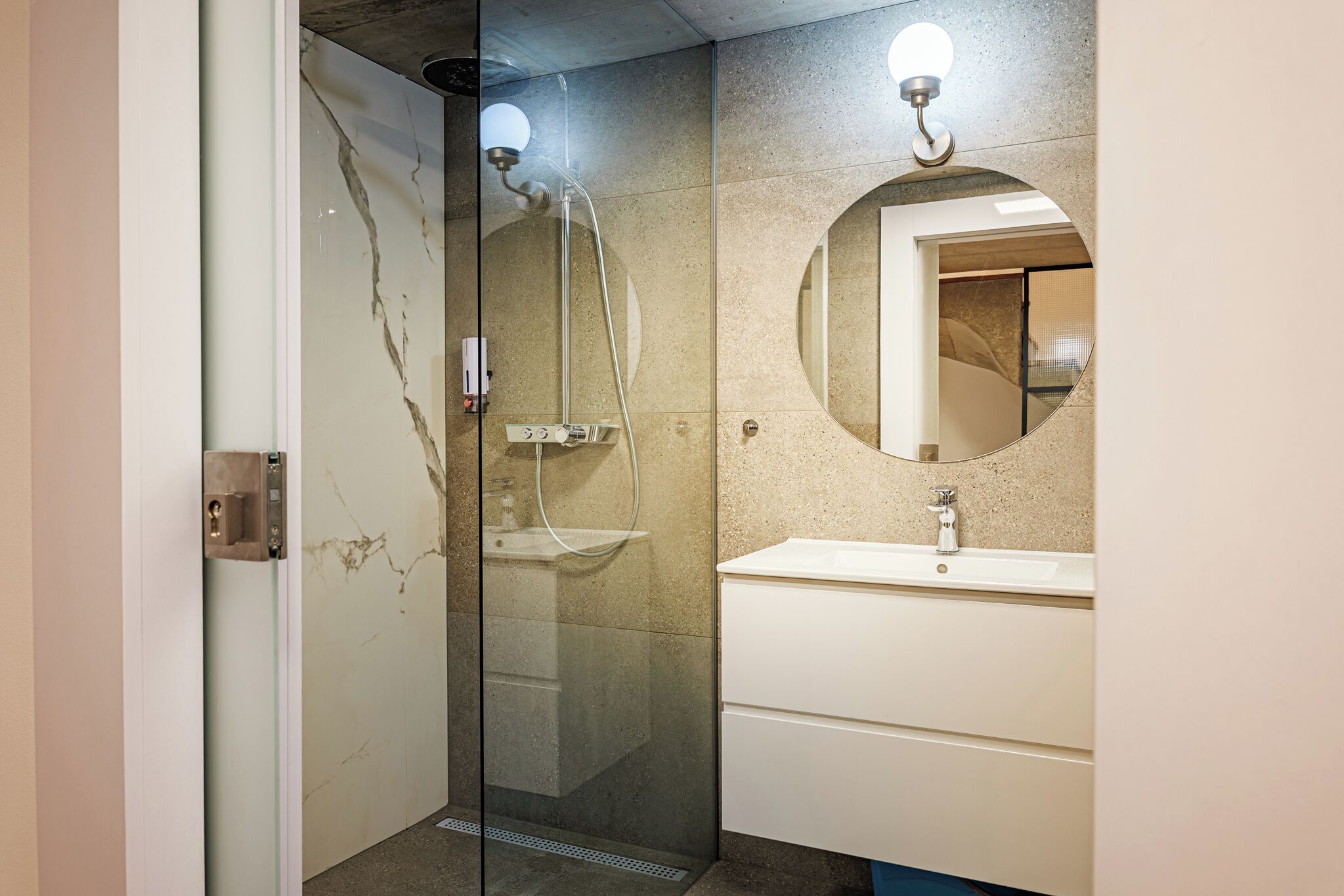 Een moderne badkamer met een douche met glazen wand, een ronde spiegel en een witte wastafel.
