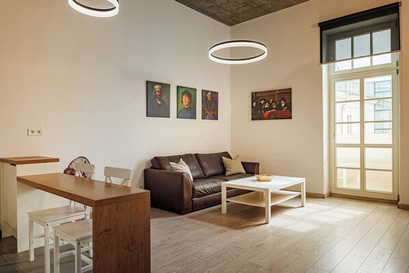 A modern living room with a brown leather sofa, white coffee table, and a dining area with a wooden table and chairs. There are three framed artworks on the wall and a large window providing natural light.