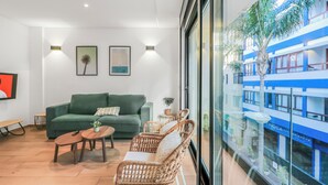 Een moderne woonkamer met een groene bank, rieten stoelen, een houten salontafel en een televisie aan de muur.