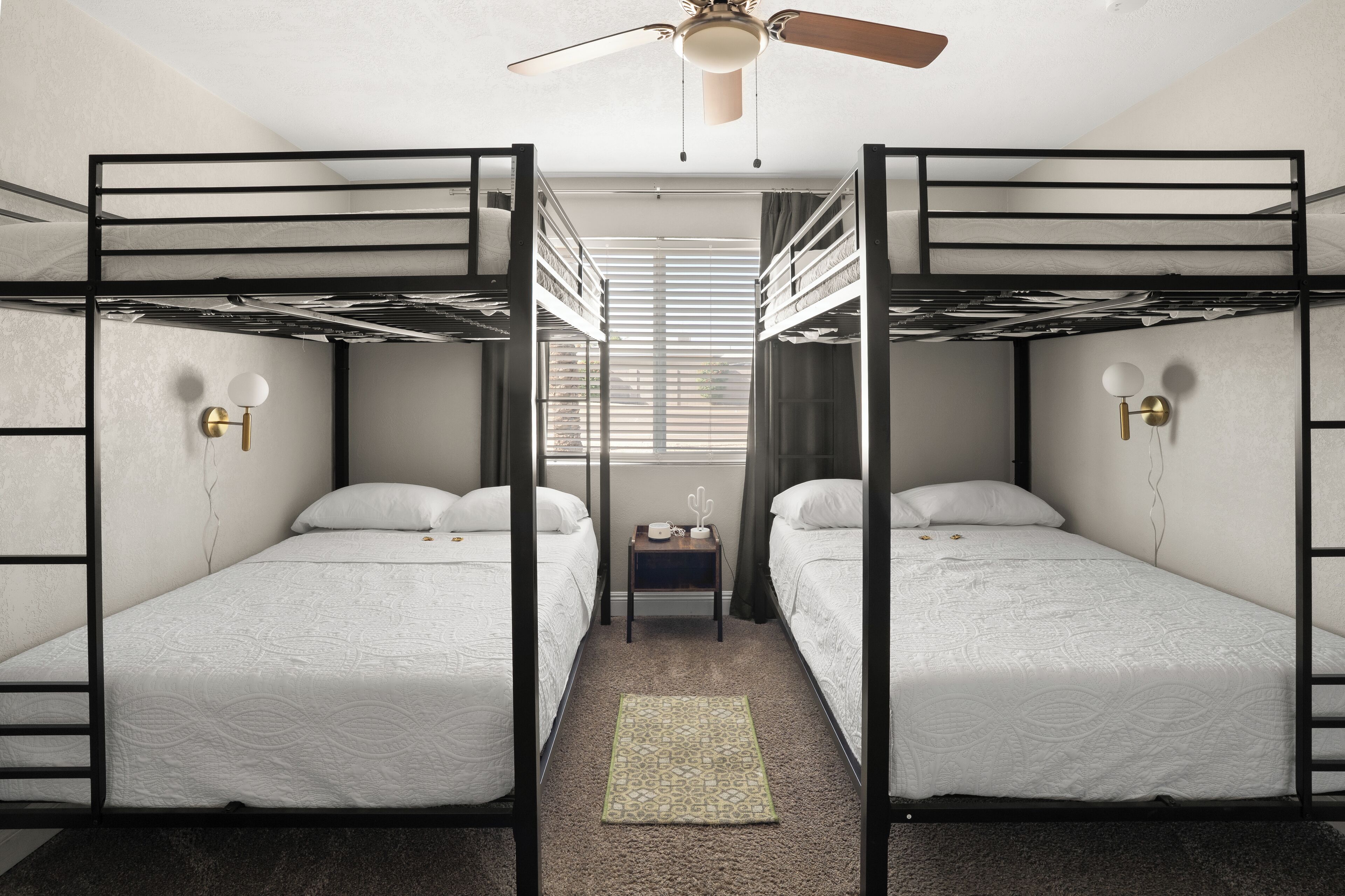 Ein Schlafsaalzimmer mit Etagenbetten, einem Deckenventilator, Nachttischlampen und einem Fenster mit Jalousien.
