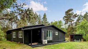 Una cabaña de troncos con un porche, un cobertizo pequeño y un patio cubierto de césped rodeado de árboles.