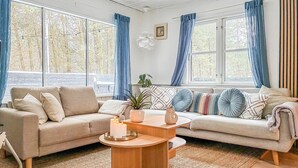 Una sala de estar acogedora con un sofá color beige, una mesa de centro de madera y un amplio ventanal con cortinas azules.